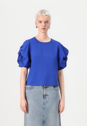 Blå kortärmad topp med texturerat tyg och puffärmar, tillsammans med en denimkjol. Toppen har rund halsringning och en avslappnad passform.
