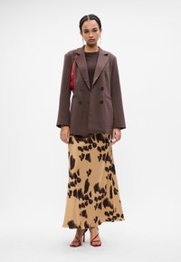 Vrouw in een bruine dubbele blazer, bruine top, beige maxirok met dierenprint, rode hoge hakken en een rode schoudertas.