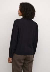 Bluză neagră cu mâneci lungi, detalii din dantelă pe umeri, decolteu rotund și manșete cu nasturi, asociată cu pantaloni maro. Textură netedă.