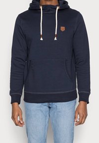 Námornicko modrá mikina s kapucí vyrobená z měkké bavlny, s klokaní kapsou, stahovacími šňůrkami, koženou nášivkou a žebrovanými manžetami a spodním lemem.