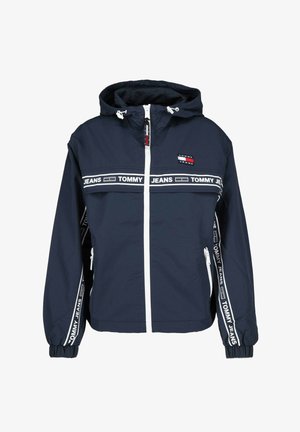Marineblauer Windbreaker aus leichtem Material, weißer Reißverschluss, verstellbare Kapuze, mit kontrastierendem weißen "TOMMY JEANS"-Schriftzug an Ärmeln und Brust.
