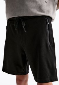 Shorts noirs en tissu texturé, dotés d'une taille à cordon de serrage et de poches latérales zippées. Longueur mi-cuisse avec une coupe décontractée.
