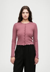 Svetlo roza rebrasti kardigan z dolgimi rokavi in okroglim ovratnikom, sprednjimi gumbi in nabranimi manšetami, v kombinaciji z visokima črnimi hlačami.