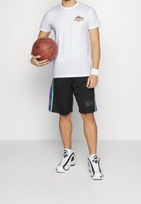 Vit t-shirt med Lakers-logotyp, svarta shorts med lila och gröna sidosträck, och vita basket skor. Person som håller en basketboll.