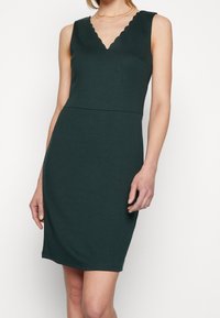 Sukienka bez rękawów w kolorze zielonym z wycięciem w kształcie litery V i falbaniastym dołem, dopasowana w talii, lekko rozkloszowana na końcu, uszyta z gładkiego, elastycznego materiału.