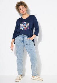 Camiseta de marina con gráfico floral "MODO CASUAL"; escote redondo, mangas tres cuartos, combinada con jeans de color azul claro y zapatillas beige.