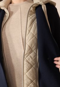 Giacca navy con fodera trapuntata crema e maglione interno beige. Presenta una chiusura con zip, tessuto testurizzato e un dettaglio con bottoni.