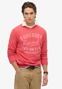 Felpa color corallo con testo "Superdry Regional Pro-Am Surf Championship", scollo rotondo e polsini a costine, abbinata a pantaloni khaki.