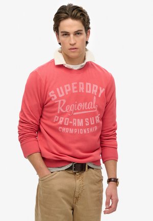 Sudadera coral con el texto "Campeonato Regional Pro-Am de Surf Superdry", cuello redondo y puños acanalados, combinada con pantalones caqui.