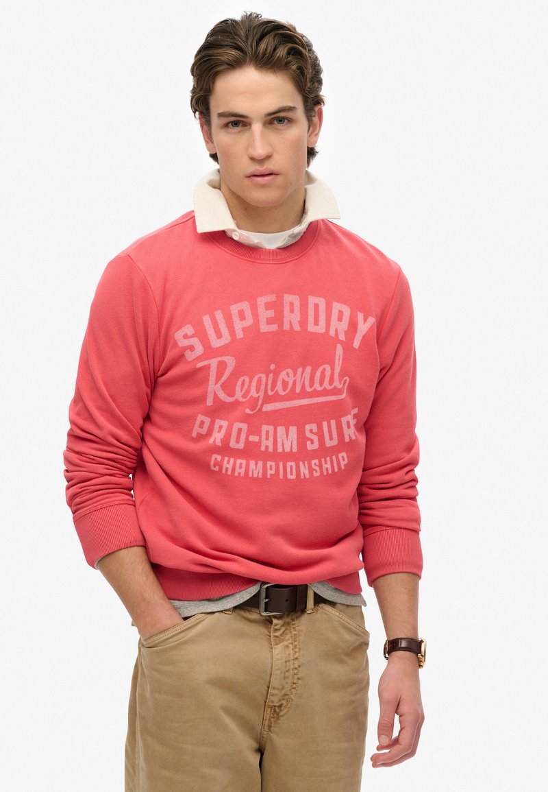 Felpa color corallo con testo "Superdry Regional Pro-Am Surf Championship", scollo rotondo e polsini a costine, abbinata a pantaloni khaki.