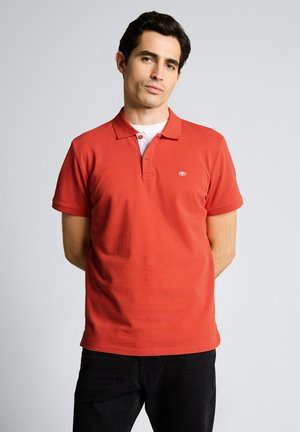 Mężczyzna o ciemnych włosach, ubrany w czerwoną koszulkę polo na białym podkoszulku, stoi ze złożonymi za plecami rękami na jednolitym szarym tle.