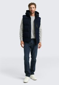 Vest puffer din material matlasat, de culoare bleumarin, cu glugă, purtat peste un hanorac gri deschis, asortat cu blugi bleumarin închis și adidași albi. Include fermoar și buzunare.
