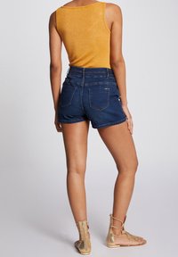 Gul, ärmlös topp med en skimrande finish, tillsammans med högmidjade mörka jeansshorts med två bakfickor. Tanfärgade sandaler med remmar.