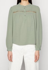 Blusa verde chiaro a maniche lunghe con scollo rotondo, chiusura a bottoni e dettagli in pizzo decorativo sul petto. Tessuto morbido con una vestibilità comoda.