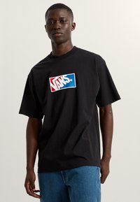 Camiseta de manga corta negra con un gran logo de Vans en rojo y azul en el pecho. La tela parece suave y tiene un corte relajado.