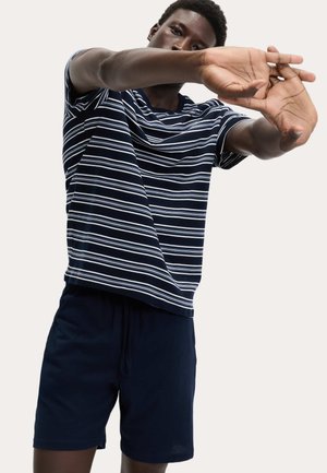 Man die een marineblauw gestreept T-shirt en marineblauwe korte broek draagt en zijn armen naar voren strekt tegen een effen lichte achtergrond.