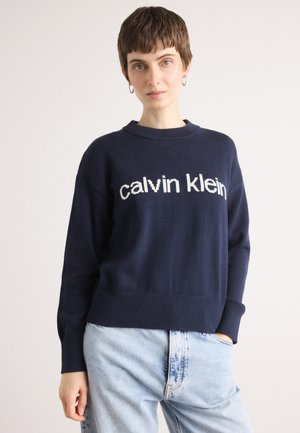 Námornícky modrý pletený sveter s rebrovaným lemom a manžetami. Na hrudi sa nachádza logo "calvin klein" v bielej farbe. Skombinovaný so svetlomodrými džínsami.
