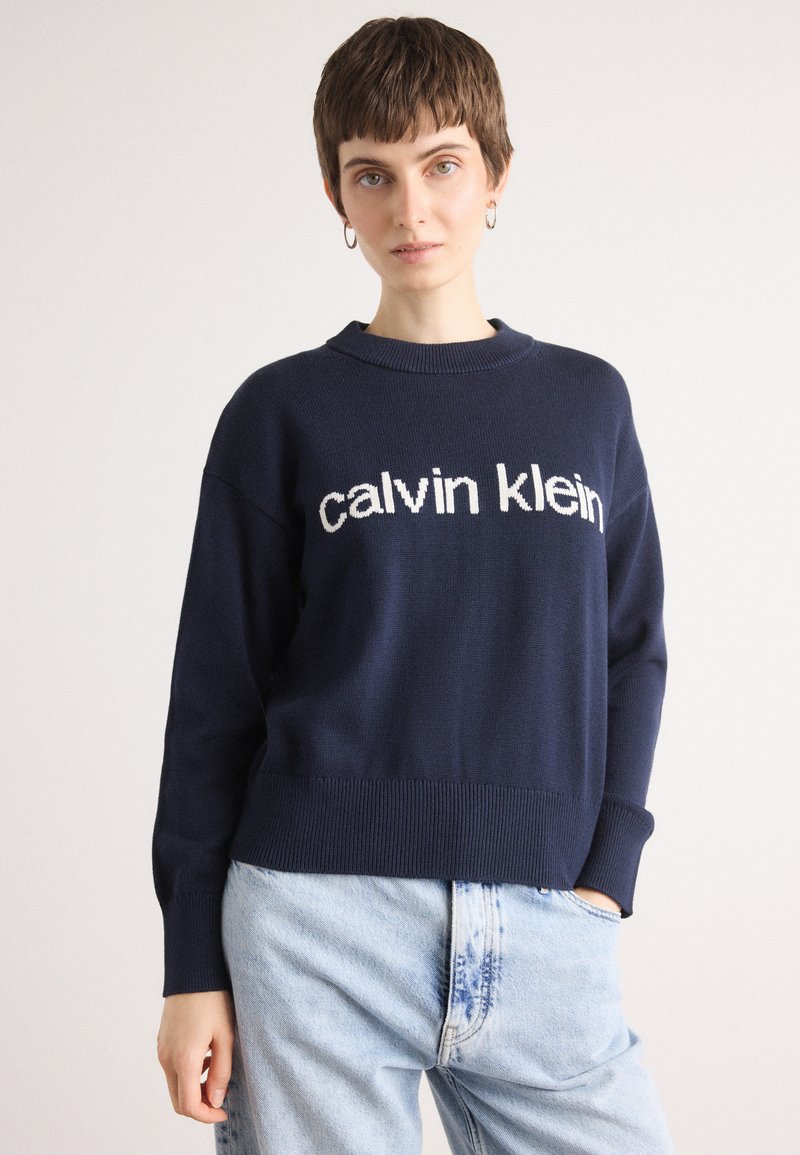 Tamsiai mėlynas nertinis megztinis su gipiūro apačia ir rankogaliais. Priekyje yra baltas "calvin klein" logotipas. Derinamas su šviesiai mėlynomis džinsomis.