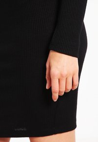 Robe noire côtelée à la coupe ajustée, manches longues, tissu texturé, longueur mi-cuisse avec un détail discret sur la couture latérale.