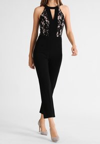 Schwarzer Jumpsuit mit einem Spitzen-Oberteil und einem Schlitz-Ausschnitt, eleganten Beinen und einer enganliegende Silhouette. Kombiniert mit hellgrauen High Heels.