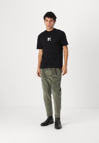 T-shirt en coton noir avec un logo blanc sur la poitrine, associé à un pantalon en velours côtelé vert olive et des bottes noires en cuir à hauteur de cheville.