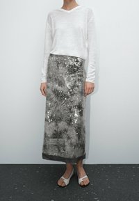 Haut blanc à manches longues associé à une longue jupe maxi grise scintillante ornée de sequins, avec une texture douce et un design plissé. Sandales blanches.