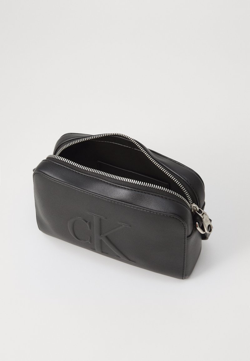 Pochette en cuir noir avec un logo "CK" en relief, fermeture éclair et matériel en ton argent. Forme rectangulaire compacte avec une texture lisse.