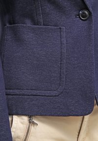 Granatowa kurtka z teksturowanej tkaniny, zapięcie na pojedynczy guzik i naszywana kieszeń po lewej stronie. Nosi się ją na jasne spodnie.