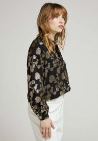 Blouse noire avec un motif floral doré, manches longues bouffantes et coupe ample. Tissu texturé léger, détail de boutons au niveau de l'encolure.