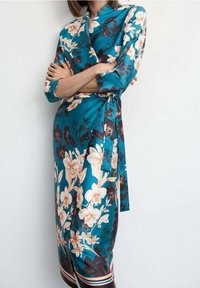 Turquoise bloemige wikkeljurk van satijn, met lange mouwen, een ceintuur in de taille en grote roomwitte en donkere bloemen op een contrasterende achtergrond.