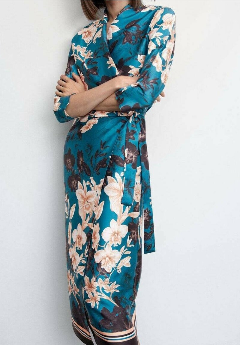 Turquoise bloemige wikkeljurk van satijn, met lange mouwen, een ceintuur in de taille en grote roomwitte en donkere bloemen op een contrasterende achtergrond.