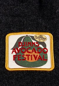 Tela de punto negra con un parche rectangular. El parche tiene un gráfico de aguacate verde, texto rojo que dice "DJINNS AVOCADO FESTIVAL" y un borde amarillo.