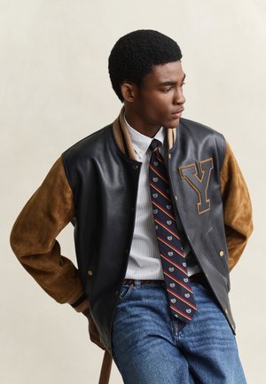 Jeune homme portant une veste varsity noire et marron avec un grand patch "Y", une cravate rayée, une chemise blanche et un jean bleu, regardant sur le côté.