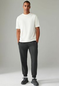 Witte katoenen T-shirt met korte mouwen, gecombineerd met donkergrijze joggingbroek met enkelboorden en zijzakken; zwarte sportieve schoenen.