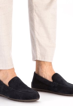 Zwarte suede instapschoenen met een ronde neus en decoratieve stiksels, gecombineerd met lichtbeige geruite broeken. Platter rubberen zool zichtbaar.
