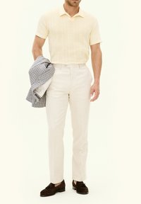 Ljust gult stickat poloärmad skjorta, krämfärgade skräddarsydda byxor och mörkbruna mockaskor, håller en rutigt grå kavaj.