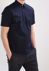 Polo-shirt à manches courtes navy avec patte de boutonnage et poignets rabattus. Le tissu a une texture lisse et une silhouette ajustée.
