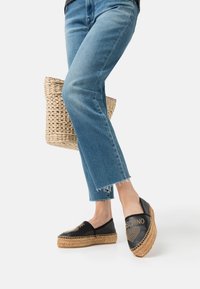 Black leather espadrilles with woven jute soles, featuring a studded heart design and the brand name. Worn with frayed denim jeans and a straw bag.