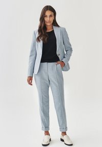 Lichtblauwe op maat gemaakte blazer met een enkele knop, gecombineerd met bijpassende broek en een zwart topje. Gladde textuur met opgerolde mouwen en loafers.