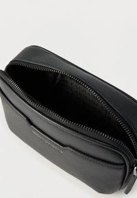 Μαύρο δερμάτινο θήκη με υφή, με ανοιχτό φερμουάρ που αποκαλύπτει το σκουρόχρωμο εσωτερικό με επαναλαμβανόμενο κείμενο του бренда και μία εμπρόσθια τσέπη.