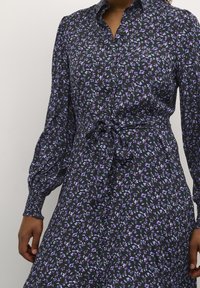 Vestito nero con bottoni, maniche lunghe, motivo floreale in viola e verde, cintura in vita e polsini plissettati. Tessuto liscio con leggero lucido.