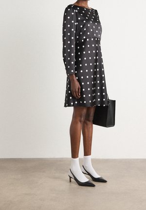 Kobieta ubrana w czarną sukienkę z długim rękawem w białe groszki, białe skarpetki, czarne szpilki, trzymająca czarną torebkę, stojąca na szarej podłodze.