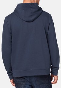 Granatowy hoodie wykonany z miękkiej mieszanki bawełny. Posiada kieszeń kangurzą, ściągacze na mankietach oraz kaptur ze sznurkiem. Gładka faktura.