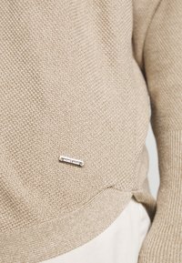 Beige stickad tröja med en strukturerad yta, som har en liten silverfärgad logotyp nära fållen och ribbade ärmslut och midja.