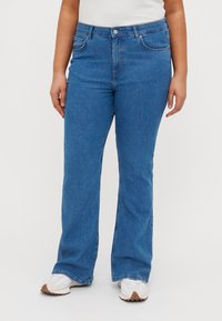 Modré rozšířené džíny vyrobené z denimu, střední pas, pět kapes a klasické zapínání na knoflík. Nosené s bílými teniskami.