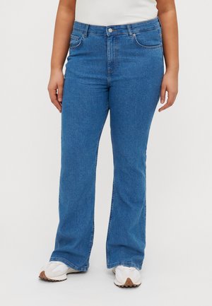 Blue flared jeans in denim fabric with a mid-rise waist, five pockets, and a classic button closure. Worn with white sneakers.