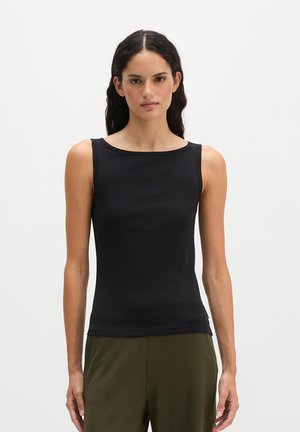 Femme aux longs cheveux foncés portant un haut noir sans manches et un pantalon vert olive, debout sur un fond clair uni.