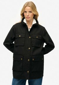 Chaqueta negra de botones con cuatro bolsillos frontales, un cuello alto y un forro interior blanco en contraste. Fabricada con tela duradera, con herrajes en tono dorado.