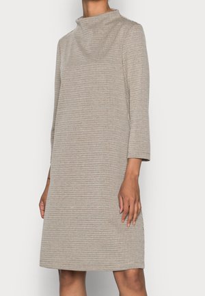 Femme portant une robe longue à manches longues beige et noire à carreaux, longueur genou, avec un col montant, debout sur un fond uni.