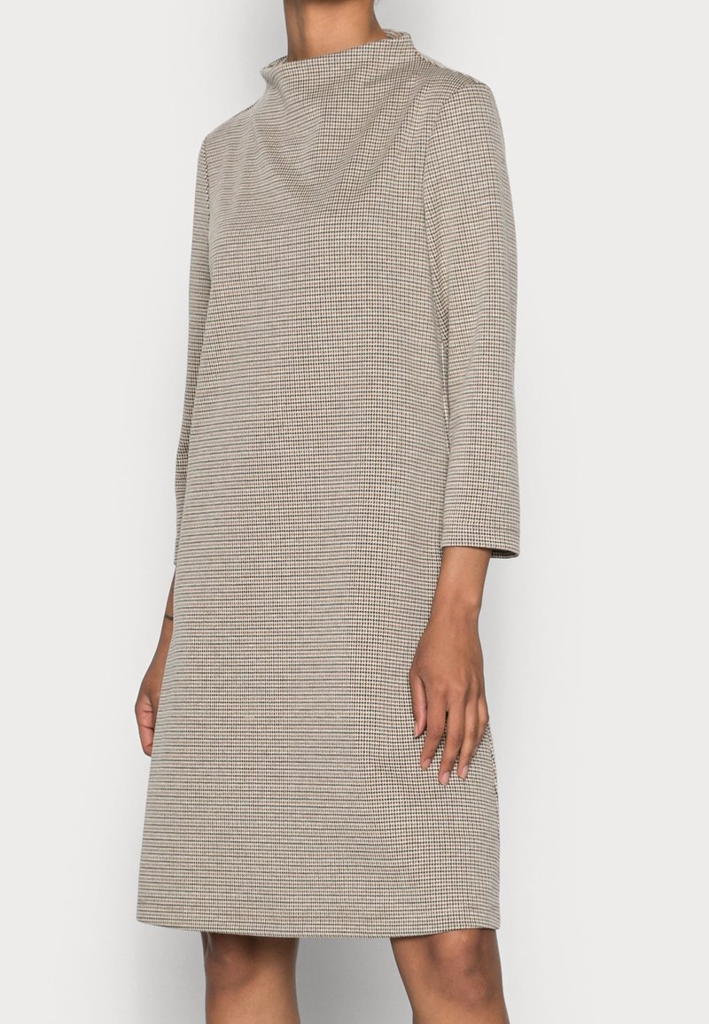 Femme portant une robe longue à manches longues beige et noire à carreaux, longueur genou, avec un col montant, debout sur un fond uni.